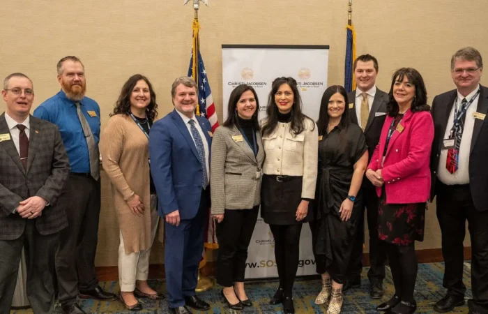 Secretary Christi Jacobsen: 'Montana further protecting election integrity, improving voter confidence by piloting national program' 8 Ten professionally dressed people stand indoors in a row, smiling for a group photo in front of a banner and two flags.