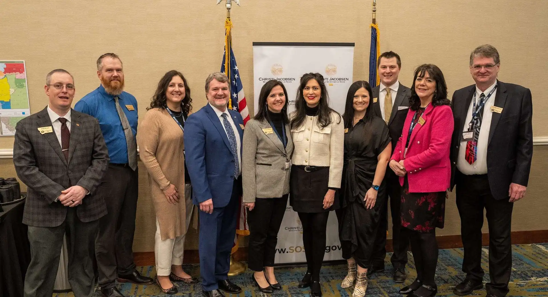 Ten professionally dressed people stand indoors in a row, smiling for a group photo in front of a banner and two flags.