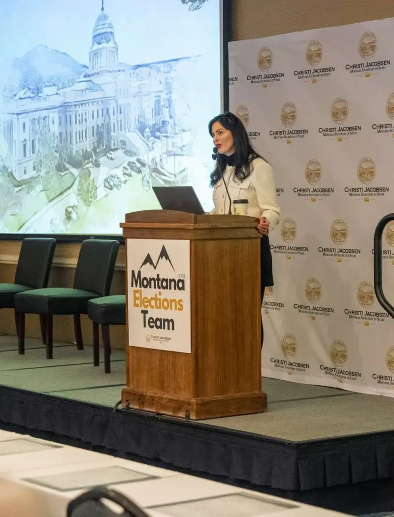 A woman, possibly Christi Jacobsen, stands at a podium labeled “Montana Elections Team” giving a presentation on absentee ballots in a conference room, with the Montana state flag and empty chairs on stage behind her. A large projected image and step-and-repeat banner are visible.