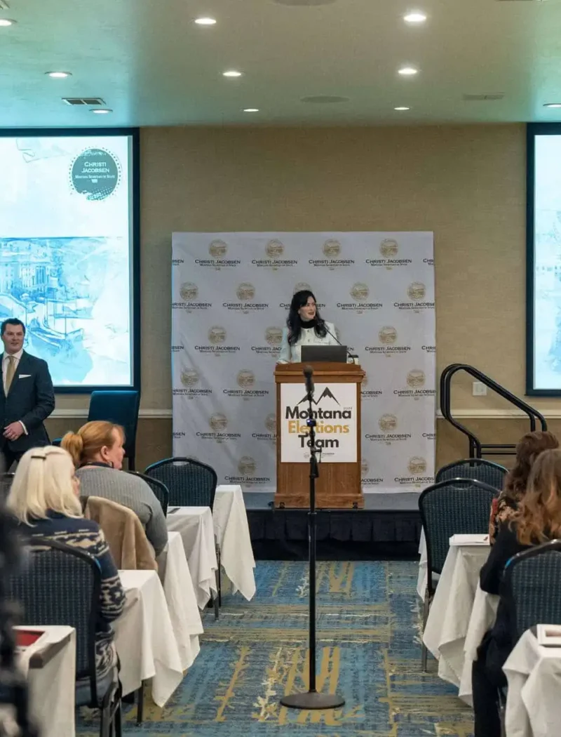Christi Jacobsen speaks at a podium labeled Montana Elections Team in a conference room, presenting an interactive absentee ballot report as attendees listen and blue-tinted images of a capitol building are projected on two large screens behind her.
