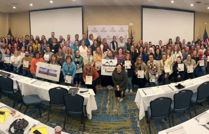 'Collaboration between Election Administrators and Secretary of State': State, local election officials praise partnership 5 A large group of election officials poses in a conference room, many holding certificates. Some individuals hold a banner reading Montana Teacher, with empty chairs and tables in the foreground. A backdrop and flags are behind the group.