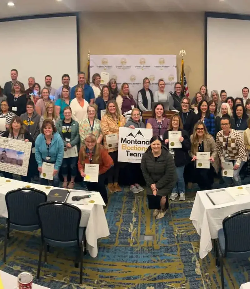 A large group of election officials poses in a conference room, many holding certificates. Some individuals hold a banner reading Montana Teacher, with empty chairs and tables in the foreground. A backdrop and flags are behind the group.