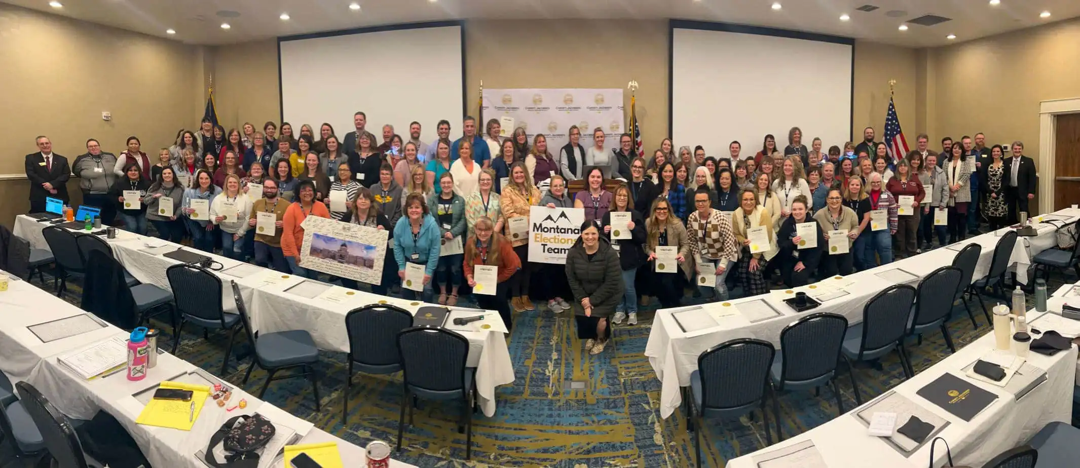 A large group of election officials poses in a conference room, many holding certificates. Some individuals hold a banner reading Montana Teacher, with empty chairs and tables in the foreground. A backdrop and flags are behind the group.