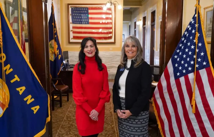 Secretary Christi Jacobsen: '2024 Candidate filing period comes to a close' 1 Two women stand smiling between Montana and U.S. flags in front of an office labeled Secretary of State. An American flag hangs on the wall inside the office. The setting appears formal and official.