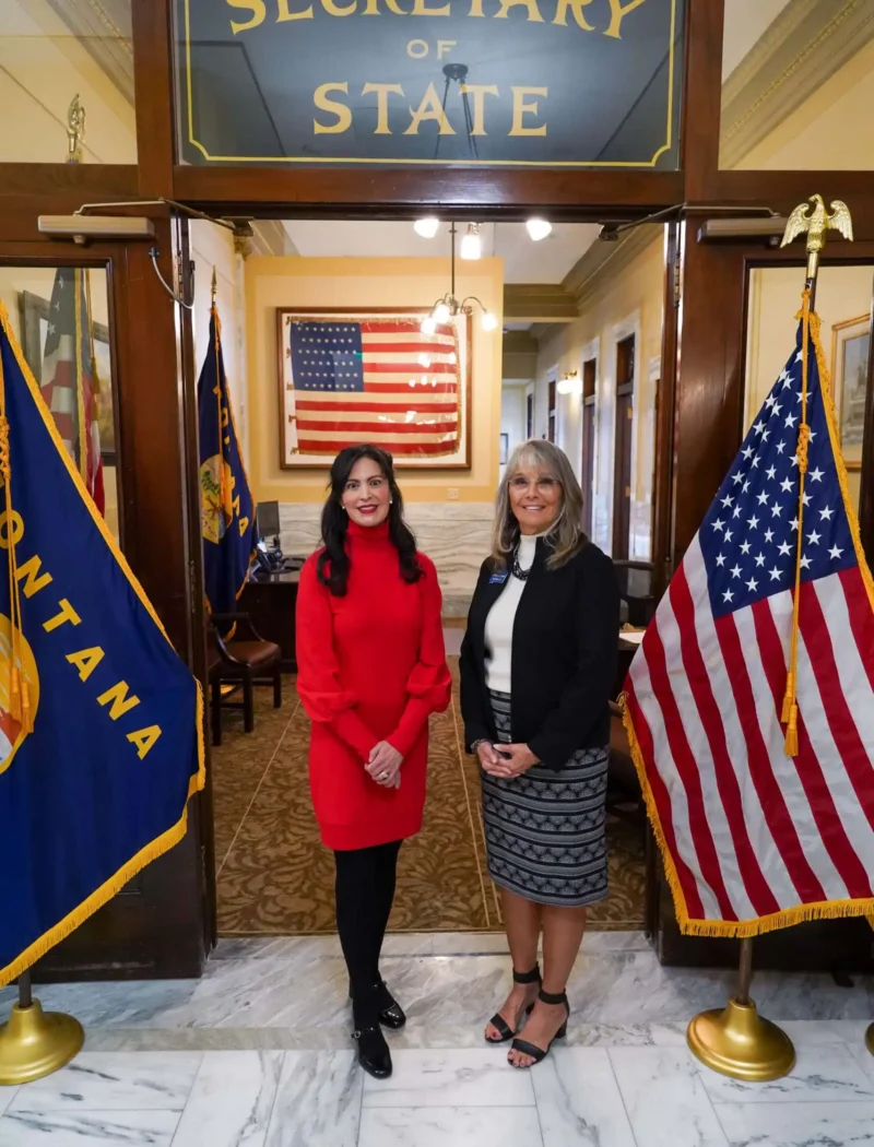 Two women stand smiling between Montana and U.S. flags in front of an office labeled Secretary of State. An American flag hangs on the wall inside the office. The setting appears formal and official.