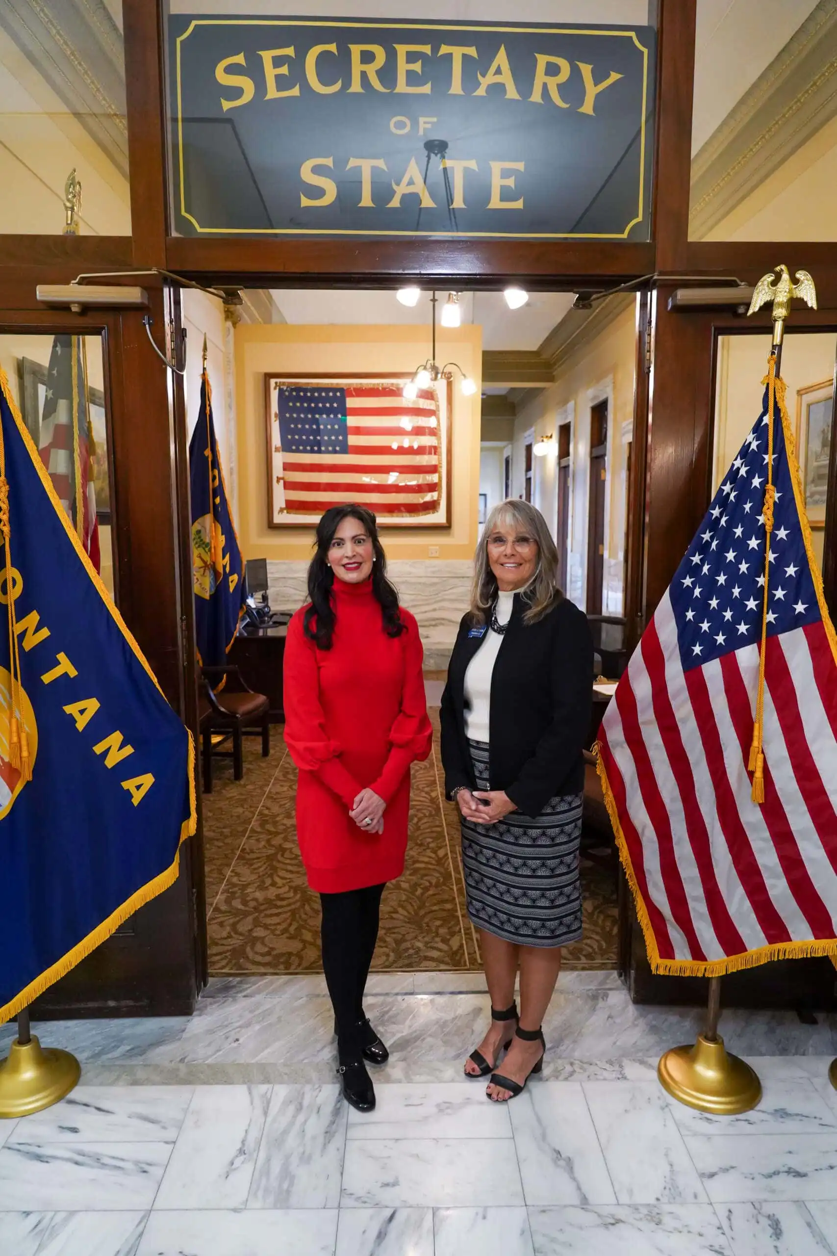 Two women stand smiling between Montana and U.S. flags in front of an office labeled Secretary of State. An American flag hangs on the wall inside the office. The setting appears formal and official.