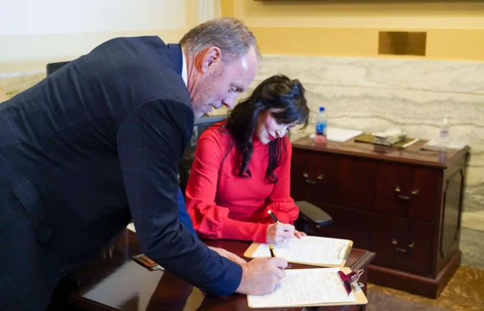 Secretary Christi Jacobsen: 'Congressional candidates highlight the week of candidate filing' 8 A man and a woman sign documents at a wooden desk in an office. The woman, wearing a red blouse, is seated, while the man in a checked suit jacket stands beside her, both focused on the paperwork.