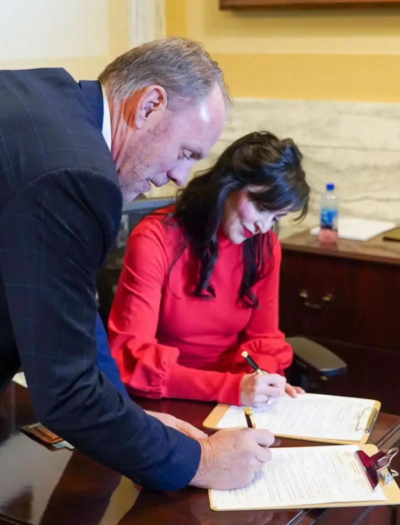 A man and a woman sign documents at a wooden desk in an office. The woman, wearing a red blouse, is seated, while the man in a checked suit jacket stands beside her, both focused on the paperwork.