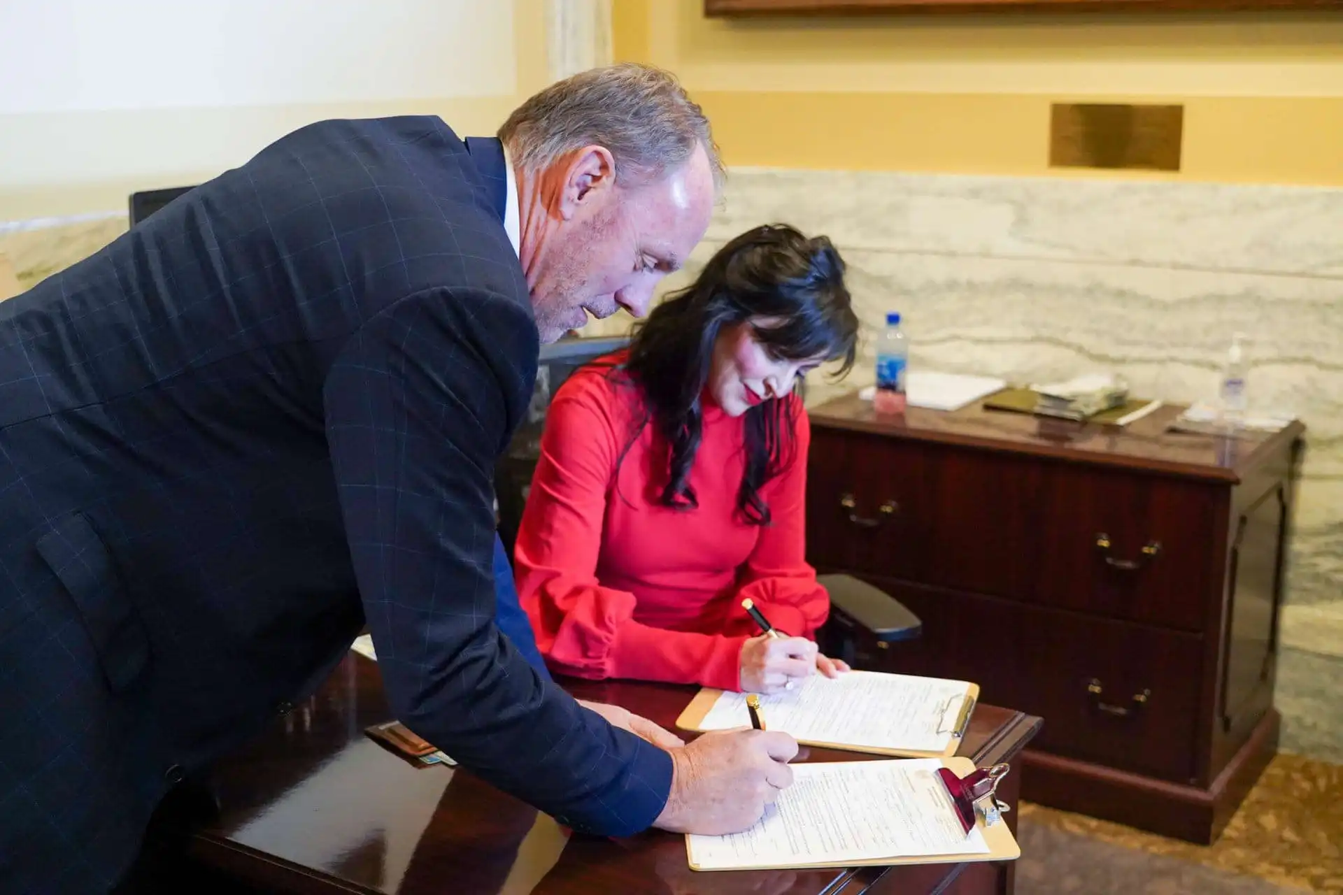 A man and a woman sign documents at a wooden desk in an office. The woman, wearing a red blouse, is seated, while the man in a checked suit jacket stands beside her, both focused on the paperwork.