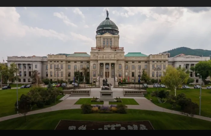 A large, domed government building with columns and a statue in front, surrounded by green lawns, trees, and parked cars—a distinctive scene from 2021 as new Montana businesses emerged against hills and a cloudy sky backdrop.