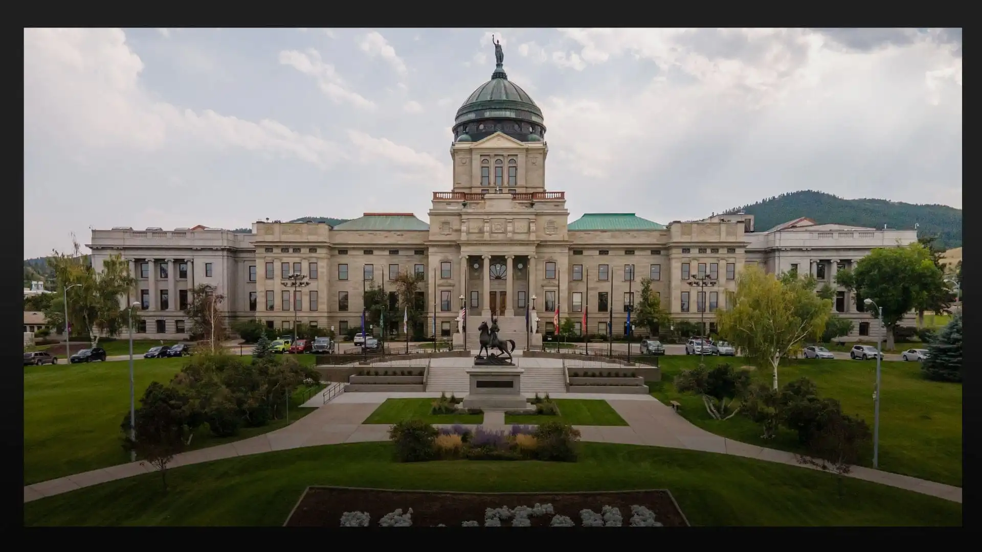 A large, domed government building with columns and a statue in front, surrounded by green lawns, trees, and parked cars—a distinctive scene from 2021 as new Montana businesses emerged against hills and a cloudy sky backdrop.