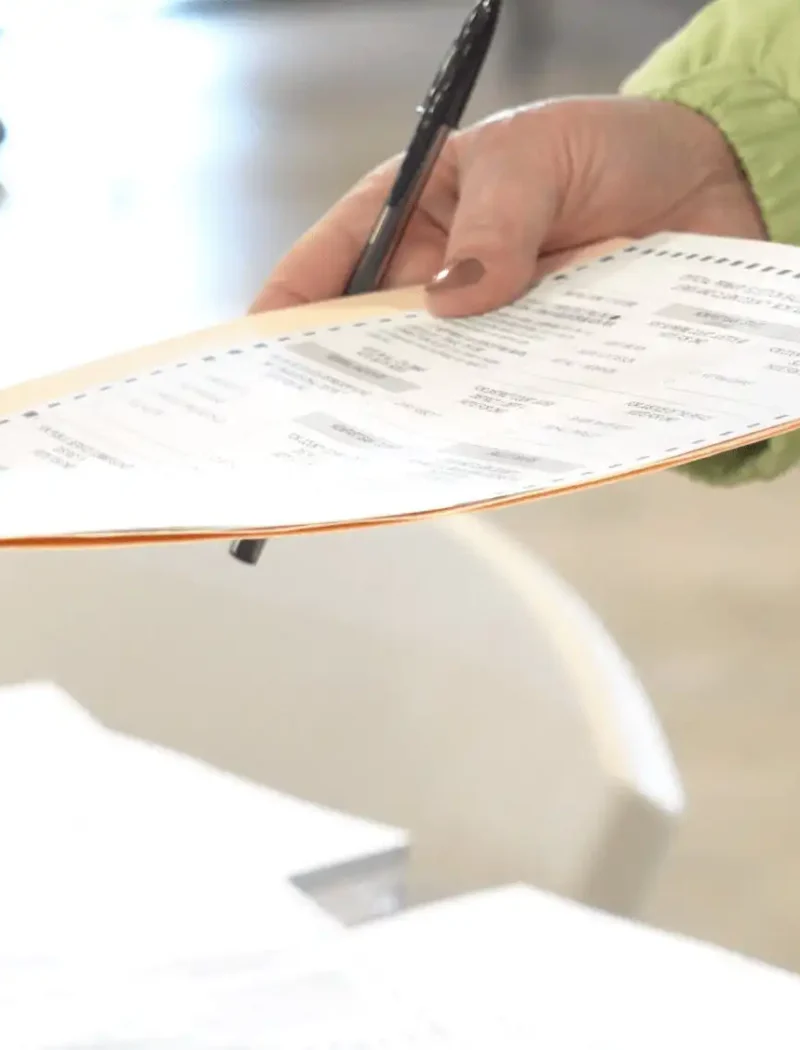 Montana Secretary of State Christi Jacobsen certifies 2022 General Election ballot 5 Close-up of a person handing a paper ballot to another at a polling place, with 2022 General Election documents and ballot certification materials from the Montana Secretary of State visible in the background.