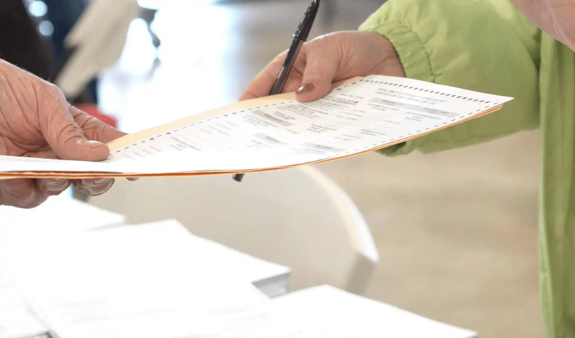 Close-up of a person handing a paper ballot to another at a polling place, with 2022 General Election documents and ballot certification materials from the Montana Secretary of State visible in the background.