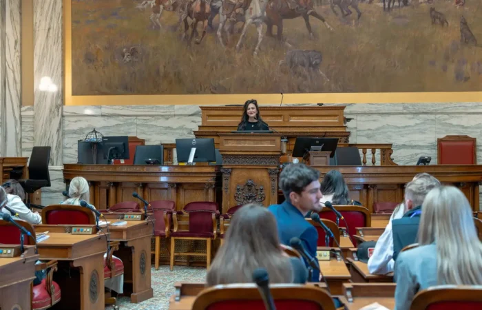Secretary Christi Jacobsen honored to kick off 2025 Montana American Legion Boys and Girls State 8 Christi Jacobsen stands at a podium in a large government chamber, addressing seated people. Behind her is a mural of horses and riders. The room has wooden desks, red chairs, and ornate decor.