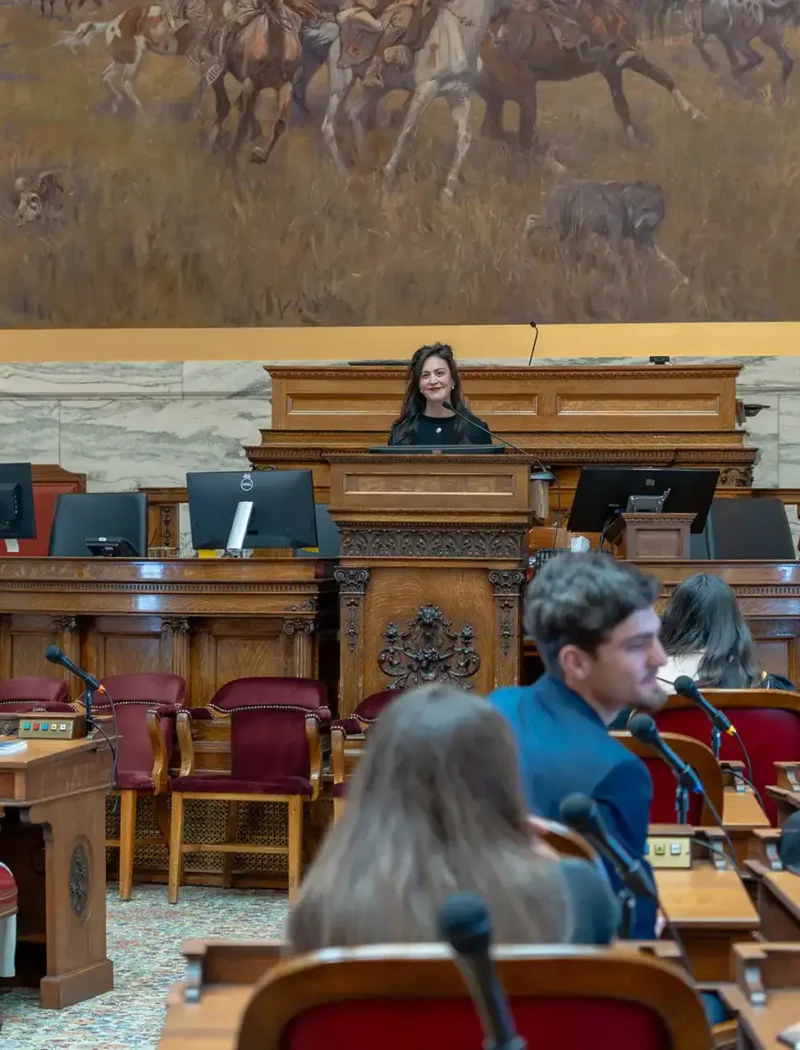 Christi Jacobsen stands at a podium in a large government chamber, addressing seated people. Behind her is a mural of horses and riders. The room has wooden desks, red chairs, and ornate decor.