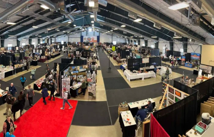 Secretary Christi Jacobsen congratulates Made in Montana businesses during 2023 Tradeshow 2 A wide view of an indoor convention center with vendor booths set up along aisles, people browsing displays, and a red carpet running through the middle of the event space.