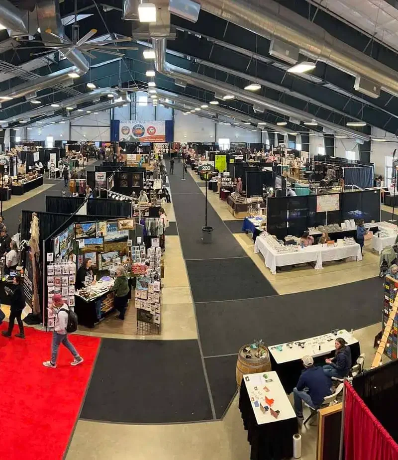 A wide view of an indoor convention center with vendor booths set up along aisles, people browsing displays, and a red carpet running through the middle of the event space.