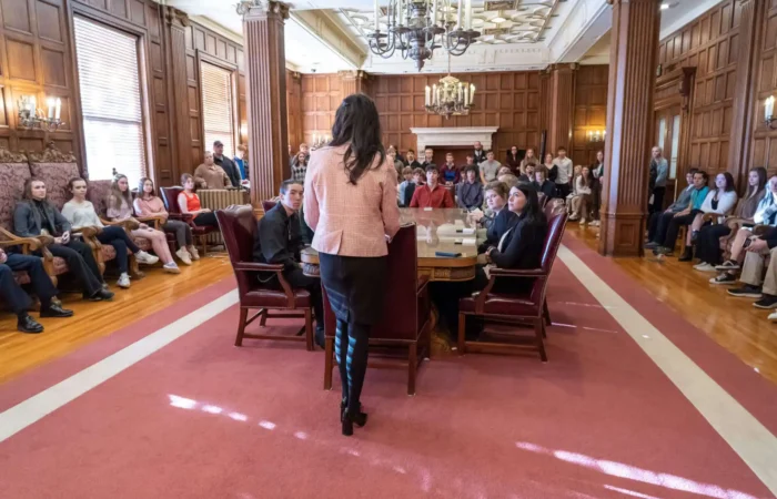 Secretary Christi Jacobsen meets with students from Cascade, Fairfield high schools 10 A woman stands addressing a room full of seated and standing people in a formal, wood-paneled meeting room with ornate chandeliers and large windows, creating an atmosphere of importance and attention.