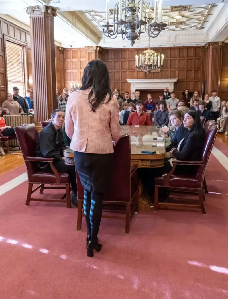 Secretary Christi Jacobsen meets with students from Cascade, Fairfield high schools 1 A woman stands addressing a room full of seated and standing people in a formal, wood-paneled meeting room with ornate chandeliers and large windows, creating an atmosphere of importance and attention.