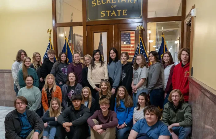 Secretary Christi Jacobsen discusses constitutions, roles of SOS Office with students from Roundup 5 A group of young people and a few adults pose for a photo in front of a Secretary of State office entrance, with American flags displayed on both sides of the doorway.