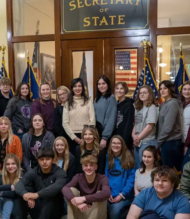 A group of young people and a few adults pose for a photo in front of a Secretary of State office entrance, with American flags displayed on both sides of the doorway.