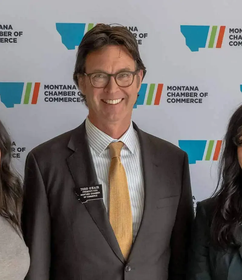 Three people in business attire, including Secretary Christi Jacobsen, stand smiling in front of a step-and-repeat banner featuring the Montana Chamber of Commerce logo. The group highlights their commitment to International Trade and business growth.