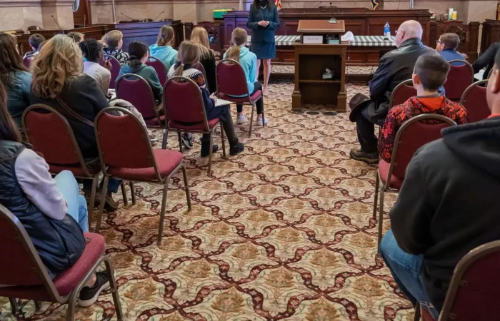 Valley Christian 6th graders visit with Secretary Christi Jacobsen, elected officials 4 A group of people, including children and adults, sit on red chairs facing a speaker at a podium in a formal room with patterned carpet and wooden desks, resembling a courtroom or government chamber.