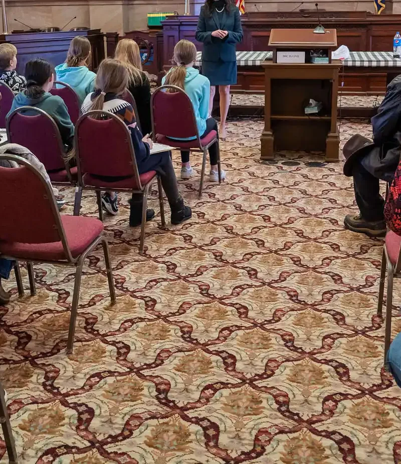 A group of people, including children and adults, sit on red chairs facing a speaker at a podium in a formal room with patterned carpet and wooden desks, resembling a courtroom or government chamber.