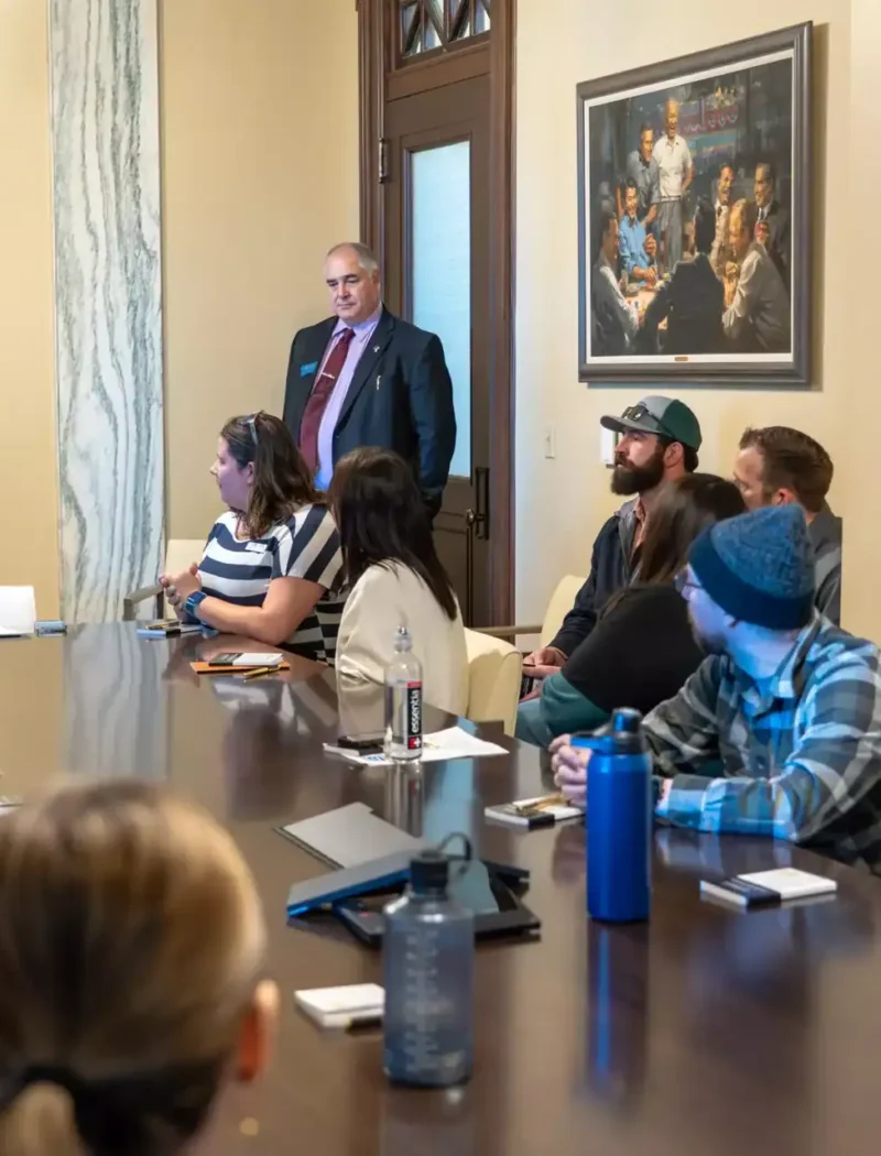 A group of western Montana business leaders sit around a conference table in a meeting room, listening to Secretary of State Christi Jacobsen speak. A man in a suit stands near the door. Laptops, notebooks, and water bottles are on the table.