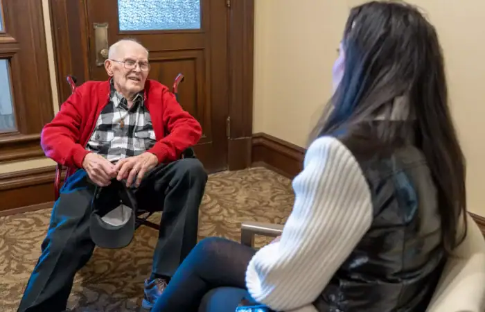 Recognizing valor: Secretary Christi Jacobsen honors 98-year-old Montana veteran Jack Kindelman 8 An elderly man in a red jacket sits and smiles while talking to a woman with long dark hair, who is wearing a white sweater and black vest, in a carpeted room near a wooden door.