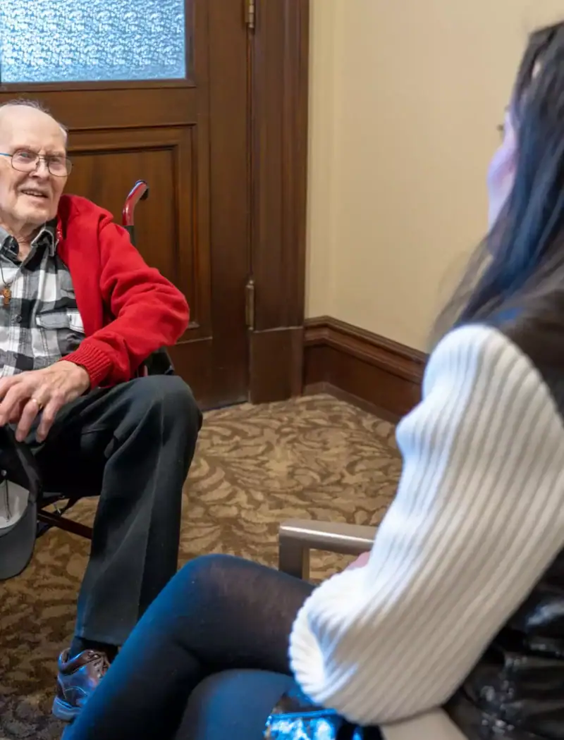 An elderly man in a red jacket sits and smiles while talking to a woman with long dark hair, who is wearing a white sweater and black vest, in a carpeted room near a wooden door.
