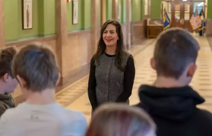 Secretary Christi Jacobsen engages with students from Corvallis, Victor at Montana State Capitol 7 A smiling woman, possibly Christi Jacobsen, wearing a black and gray checkered top, stands in a hallway with green walls and framed pictures at the Montana State Capitol, facing students whose backs are to the camera.