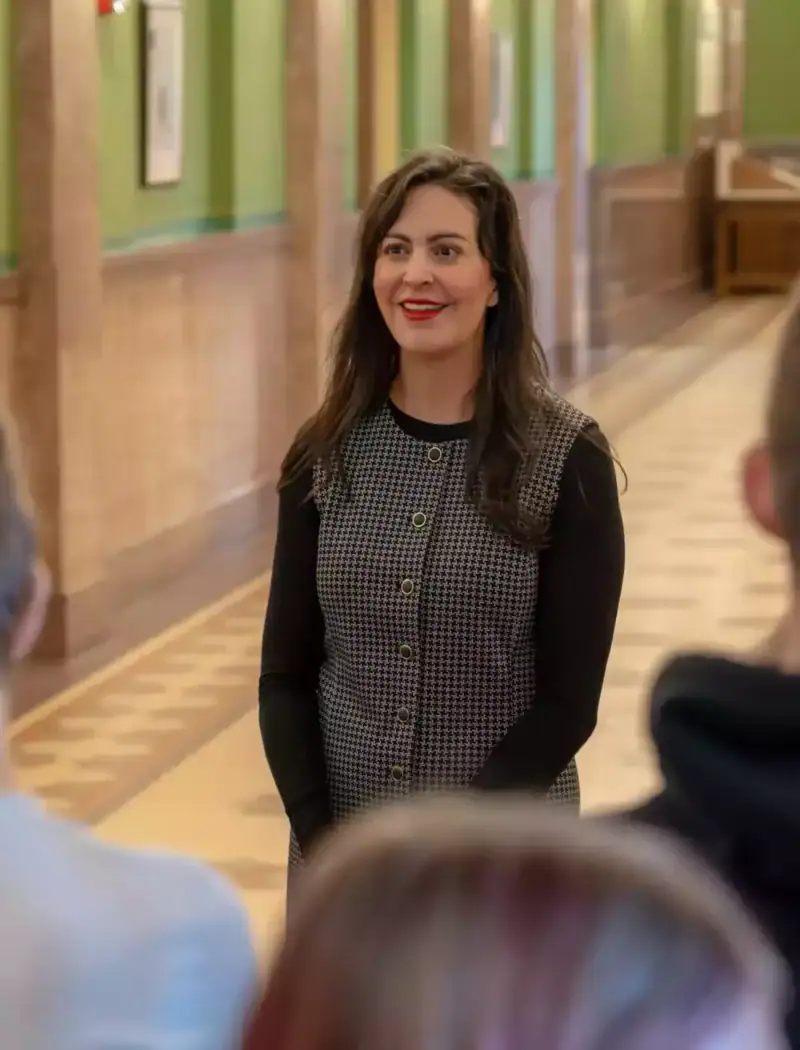 A smiling woman, possibly Christi Jacobsen, wearing a black and gray checkered top, stands in a hallway with green walls and framed pictures at the Montana State Capitol, facing students whose backs are to the camera.