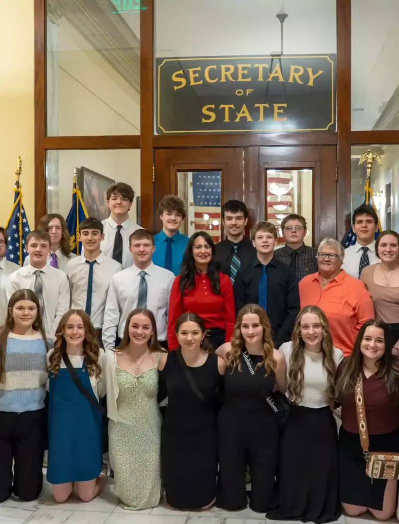 A group of young adults, including some homeschool students and adults, pose together for a photo in front of the Secretary of State office entrance inside the Montana State Capitol. Most are dressed formally and smiling at the camera.