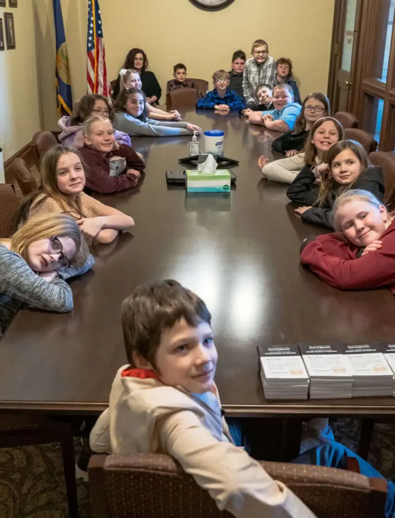 Vaughn 4th graders sit around a long conference table in a room with framed photos, large windows, and an American flag at the State Capitol. Some smile at the camera while an adult stands at the end.