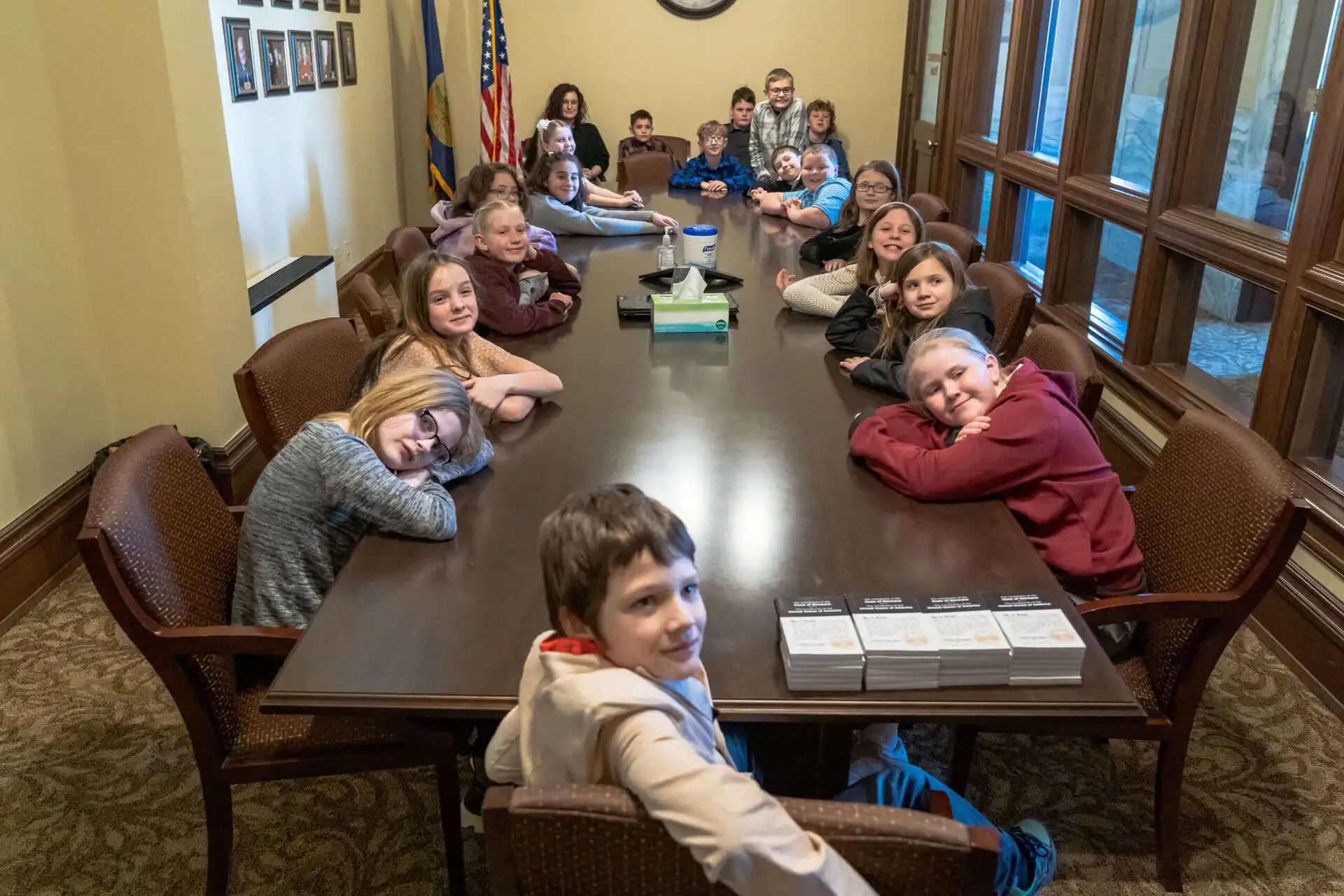 Vaughn 4th graders sit around a long conference table in a room with framed photos, large windows, and an American flag at the State Capitol. Some smile at the camera while an adult stands at the end.