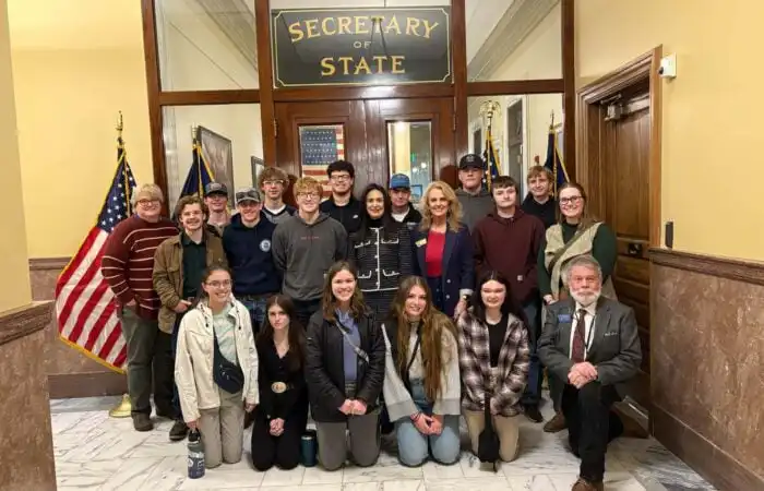 Secretary of State Christi Jacobsen engages with student groups during first half of Legislative Session 8 A group of people, including student groups, pose for a photo inside a government building in front of a “Secretary of State” sign, with American flags on either side. Most are standing, while a few are kneeling in front.