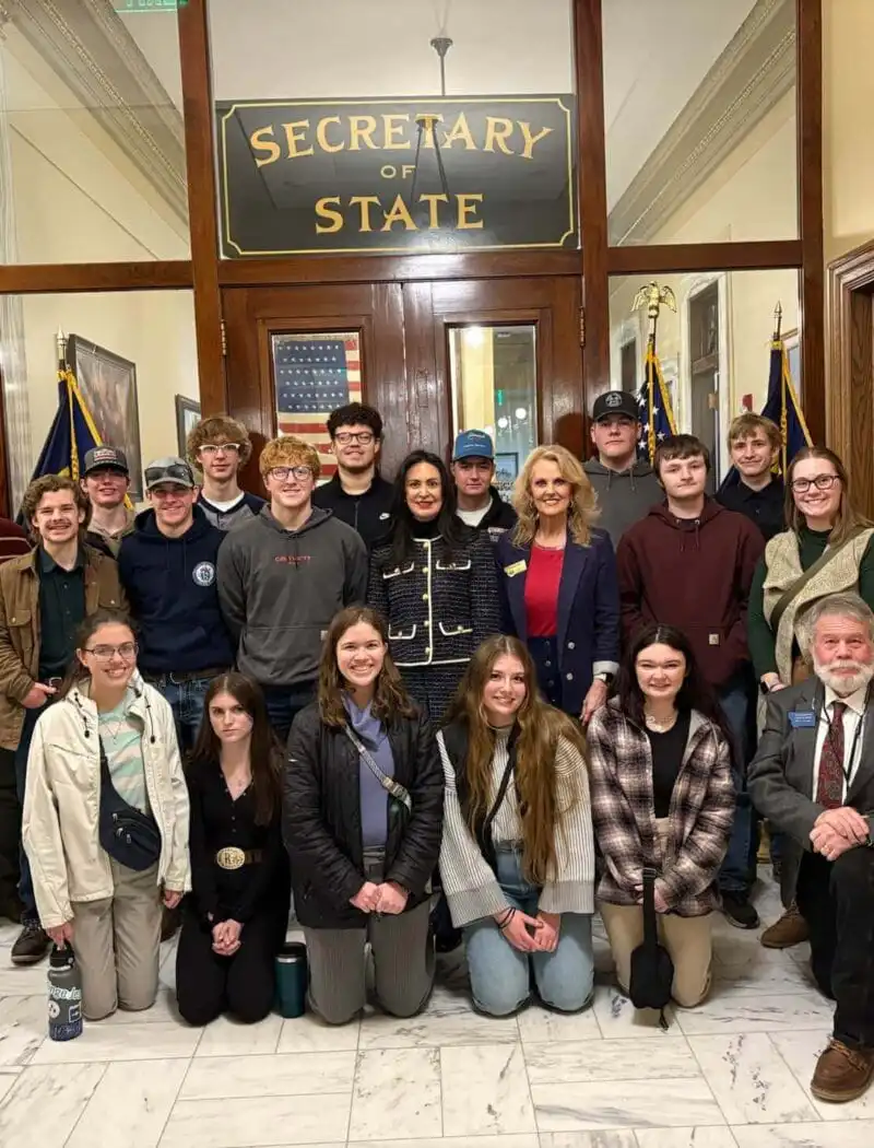 A group of people, including student groups, pose for a photo inside a government building in front of a “Secretary of State” sign, with American flags on either side. Most are standing, while a few are kneeling in front.