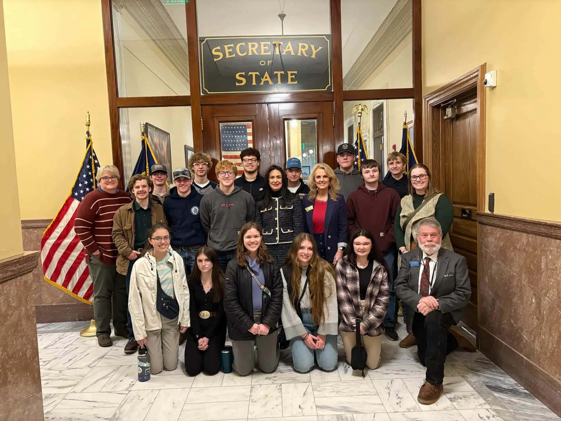 A group of people, including student groups, pose for a photo inside a government building in front of a “Secretary of State” sign, with American flags on either side. Most are standing, while a few are kneeling in front.