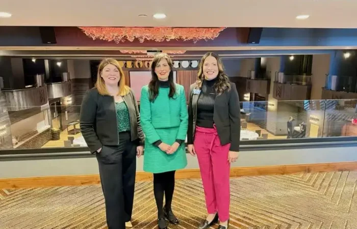 Secretary Christi Jacobsen joins esteemed colleagues at Montana Chamber's Women in Business Summit 6 Three women stand side by side in a modern, well-lit event space. The woman in the center wears a green dress, while the others wear business attire with black jackets. The background shows tables and elegant decor.