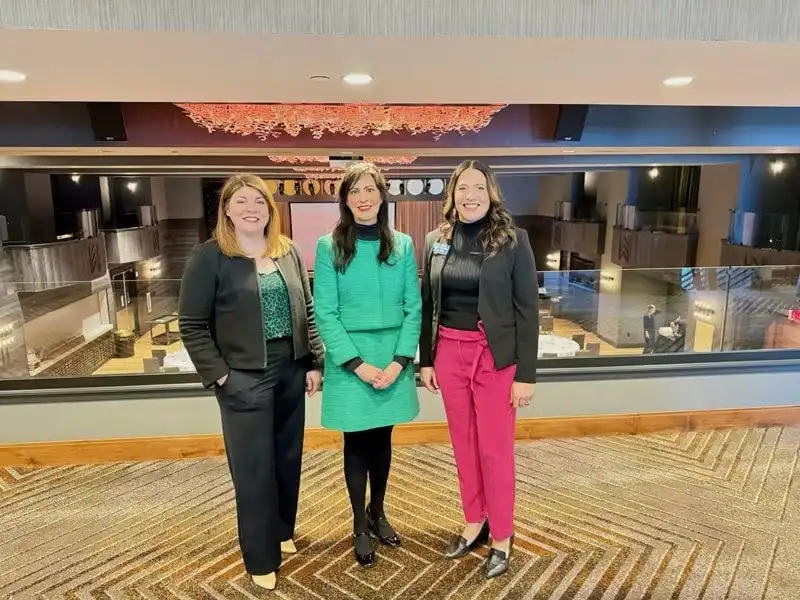 Three women stand side by side in a modern, well-lit event space. The woman in the center wears a green dress, while the others wear business attire with black jackets. The background shows tables and elegant decor.