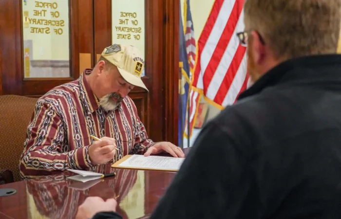 Secretary Christi Jacobsen: 'Nearly 50 new candidate filings processed this week' 2 A man wearing a Veteran hat sits at a desk signing papers, while another person sits across from him. An American flag and office doors labeled Secretary of State are visible in the background.