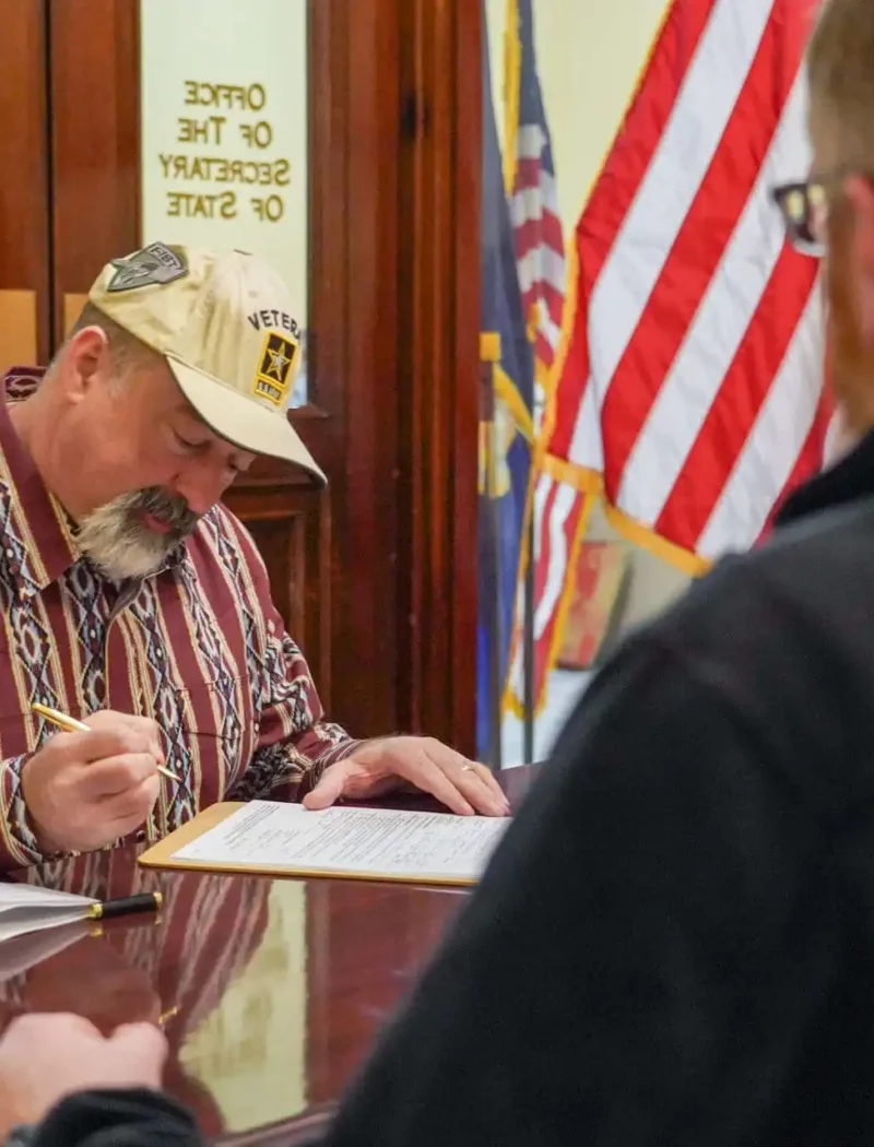 A man wearing a Veteran hat sits at a desk signing papers, while another person sits across from him. An American flag and office doors labeled Secretary of State are visible in the background.