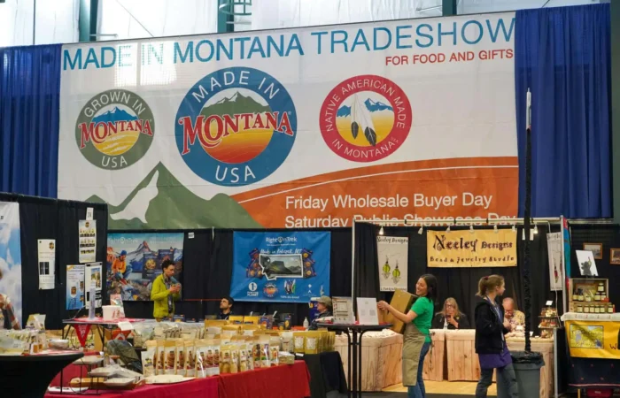 Secretary Christi Jacobsen congratulates businesses at 2024 Made in Montana Tradeshow 1 Vendors and shoppers at the Made in Montana Tradeshow, with booths selling food and gifts. Large banners display “Made in Montana” logos above the booths and announce the event details.