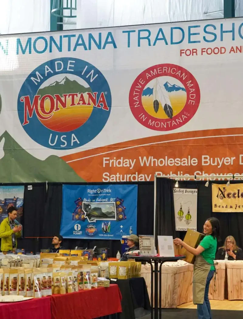 Vendors and shoppers at the Made in Montana Tradeshow, with booths selling food and gifts. Large banners display “Made in Montana” logos above the booths and announce the event details.