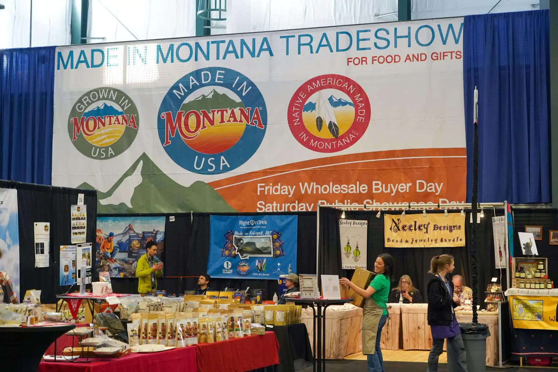 Vendors and shoppers at the Made in Montana Tradeshow, with booths selling food and gifts. Large banners display “Made in Montana” logos above the booths and announce the event details.