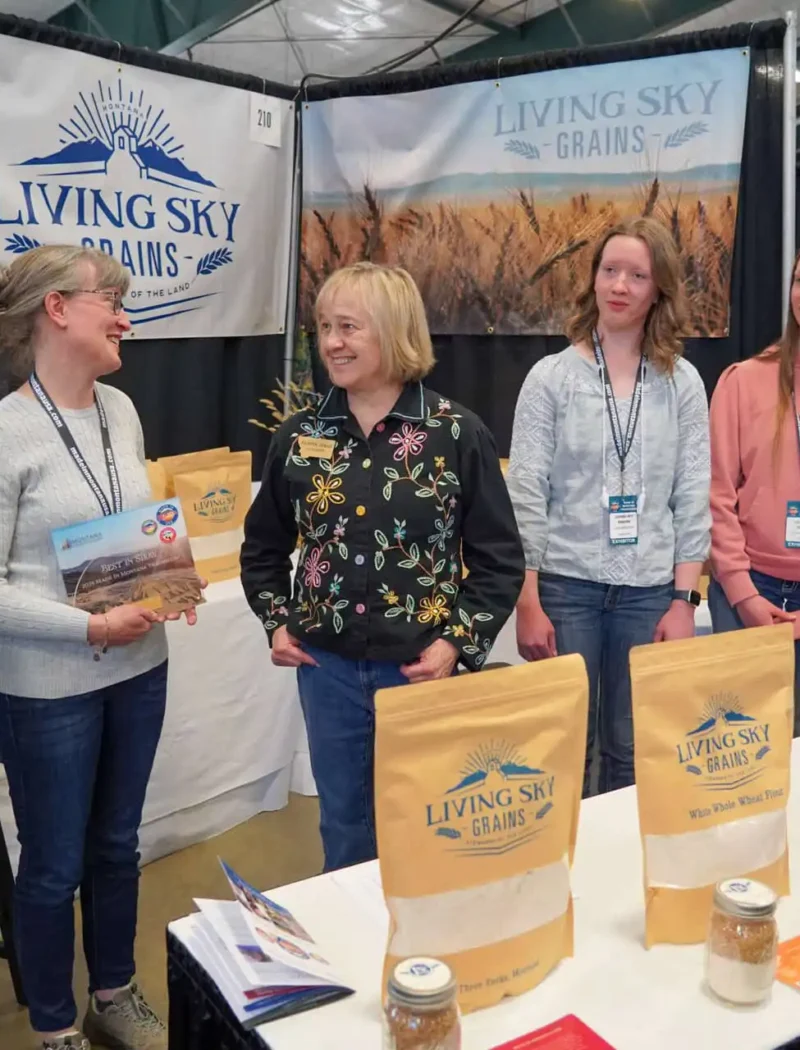 Five people stand and smile at a booth for Living Sky Grains, with various grain products displayed on the table in front of them at what appears to be an indoor market or trade show.