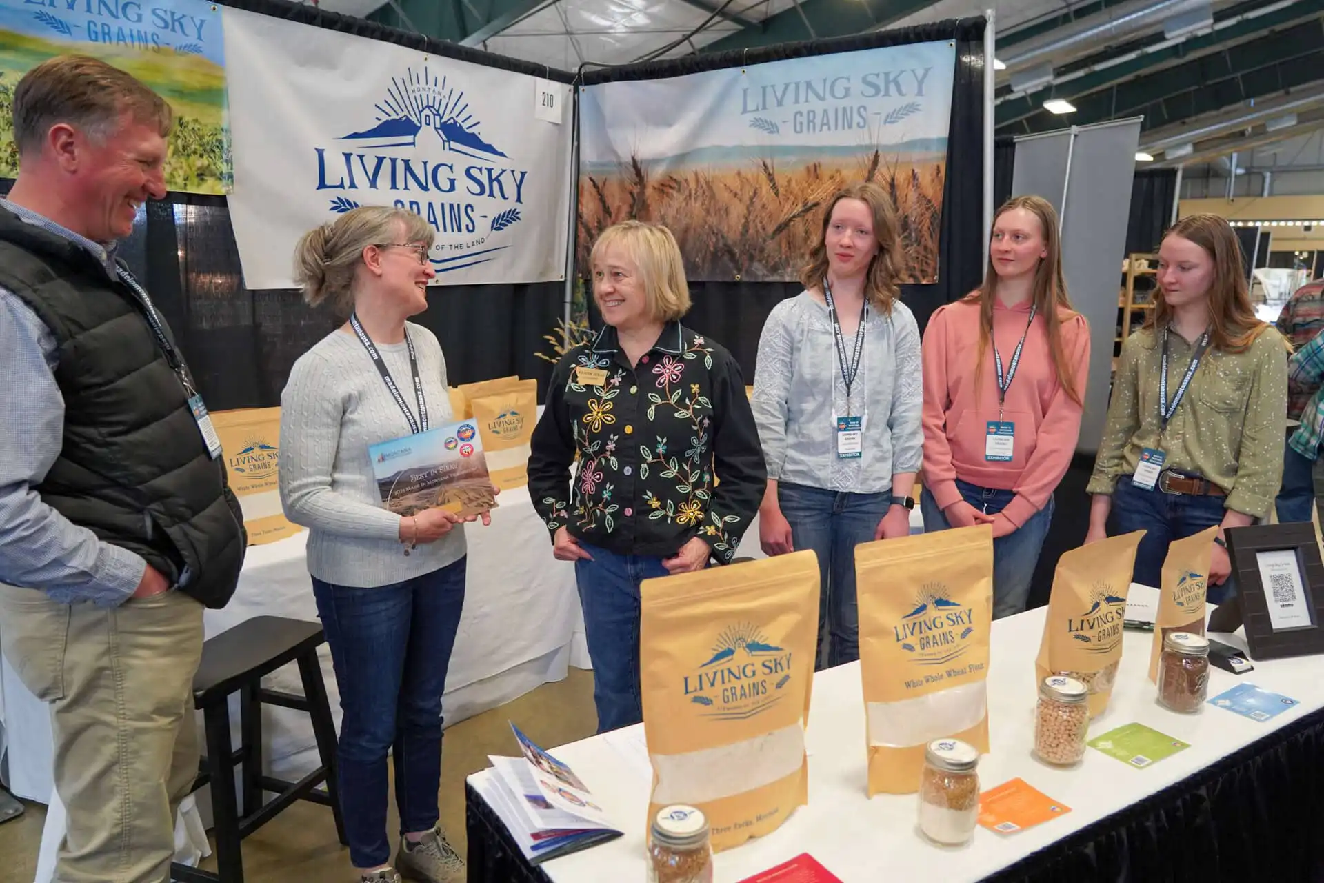 Five people stand and smile at a booth for Living Sky Grains, with various grain products displayed on the table in front of them at what appears to be an indoor market or trade show.