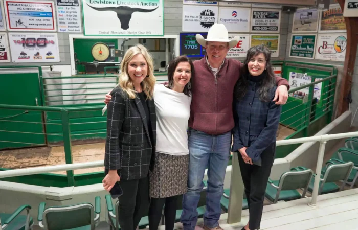 Montana Business Spotlight: Lewistown Livestock Auction a staple in central Montana 5 Four people stand smiling with arms around each other inside the Lewistown indoor livestock auction arena, with green fencing and advertisements on the walls in the background.