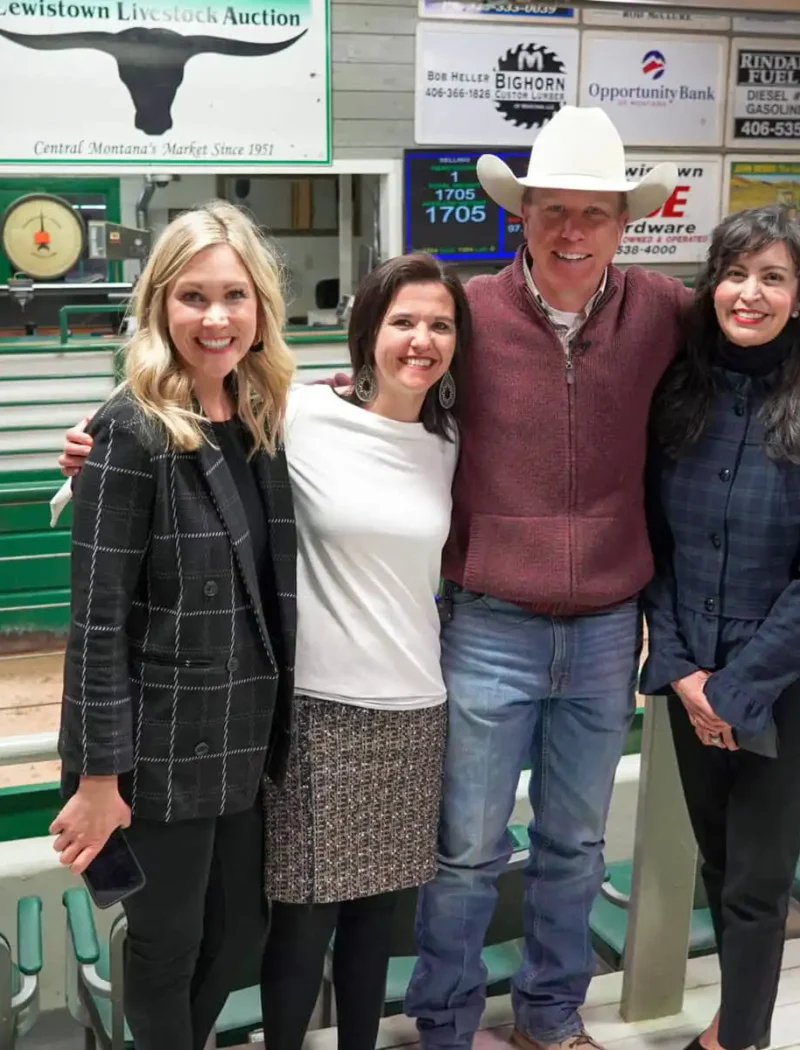 Four people stand smiling with arms around each other inside the Lewistown indoor livestock auction arena, with green fencing and advertisements on the walls in the background.